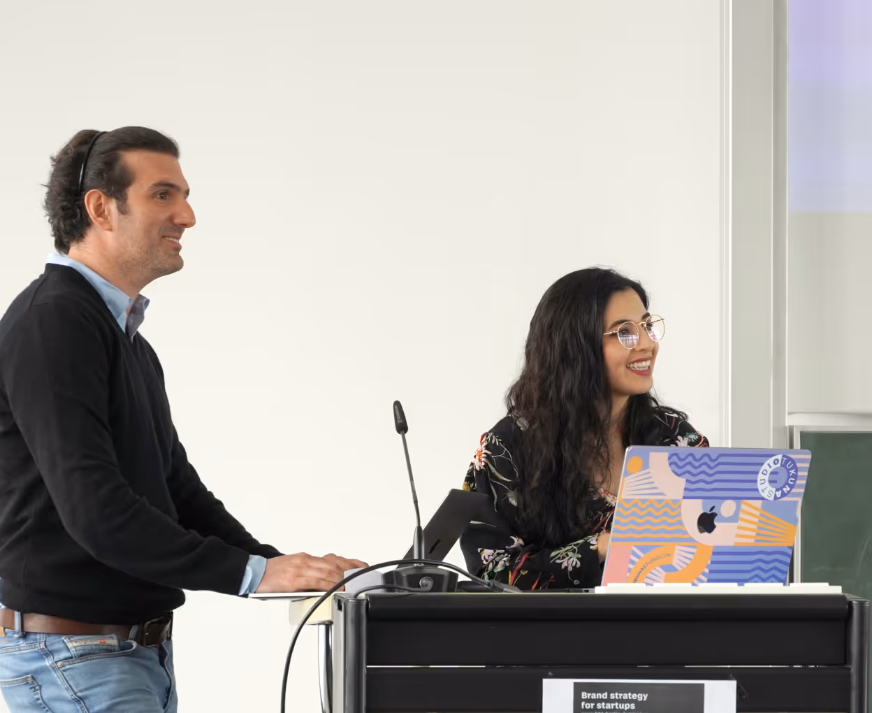 Two people at a podium, a man standing with a laptop and a smiling woman with glasses and a colorful laptop.