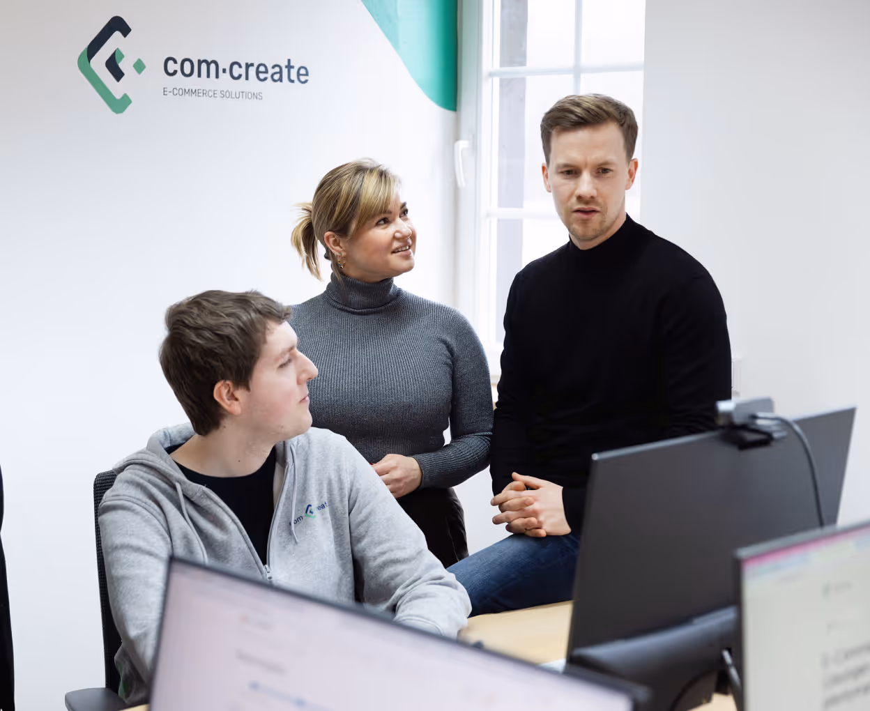 Three colleagues discussing work in an office with computer monitors and a Com-Create e-commerce solutions logo on the wall.