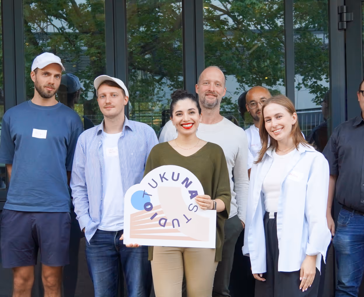 Group of six diverse adults smiling, standing outdoors in front of glass doors, one woman holding a sign that reads 'TUKUNA STUDIO.'