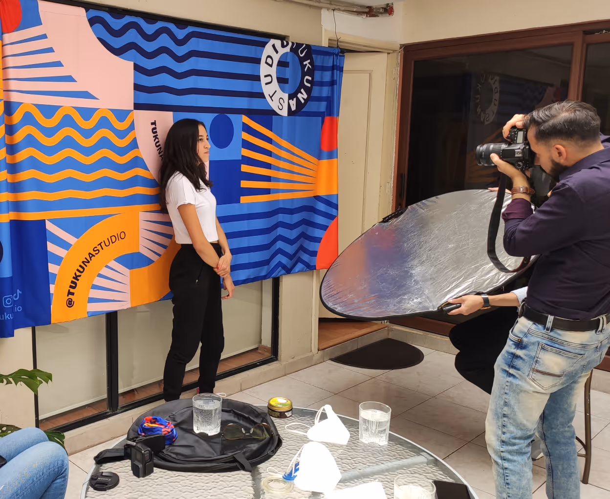 Four people in a bright room with tiled floor, one holding a reflector, one wearing a mask, and one holding a camera near a glass table with photography gear.
