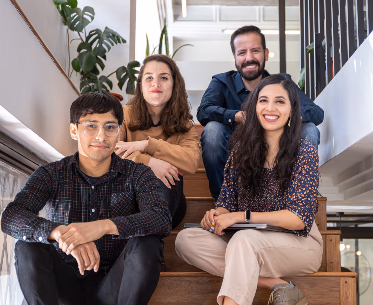 Four diverse young adults sitting on wooden stairs inside a modern office, smiling and looking at the camera.