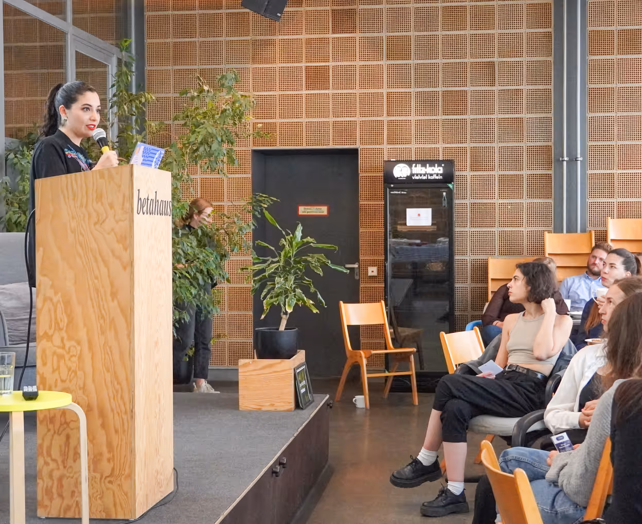 Woman speaking at a wooden podium labeled 'betahaus' to an audience seated in a modern room with plants and wood panel walls.