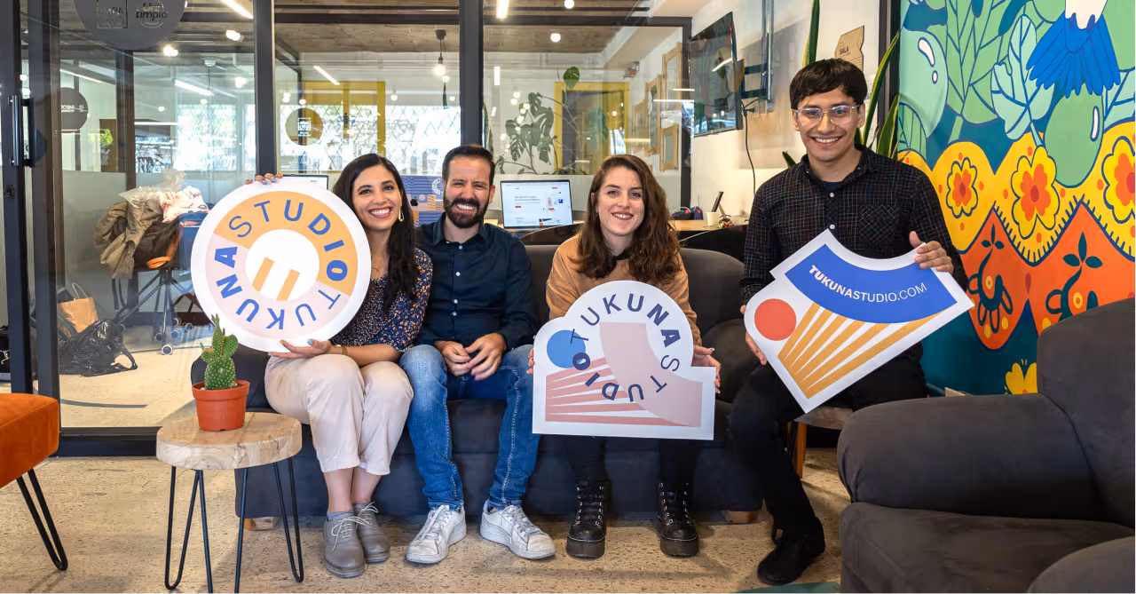 Four smiling people sitting on a couch in a modern office, each holding a colorful sign with 'TUKUNA STUDIO' text.
