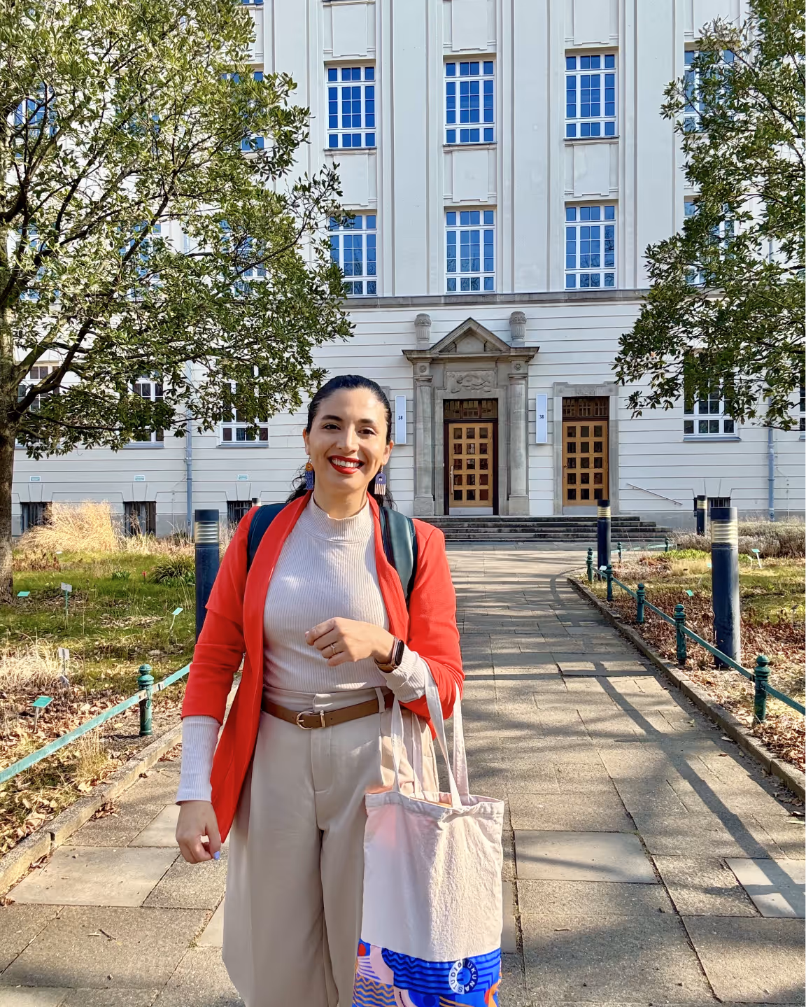 Smiling woman wearing a red blazer and beige pants standing on a walkway in front of a white building with trees on either side.
