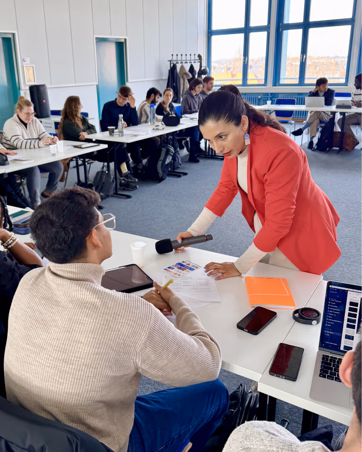 Woman in red blazer holding a microphone and leaning over a table while talking to a seated man wearing glasses and a beige sweater in a classroom setting.