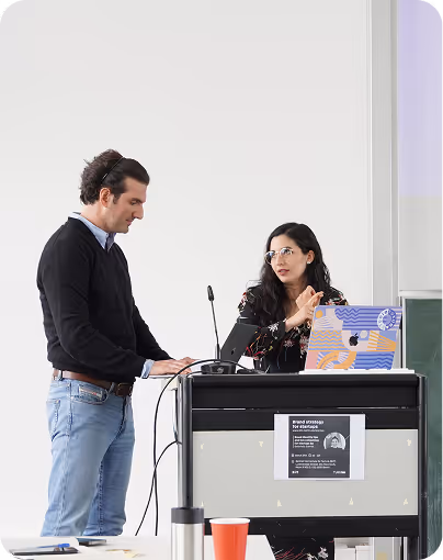 Two people standing at a podium with a laptop, engaged in discussion or presentation.
