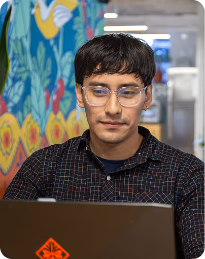 Young man wearing glasses working on a laptop with a colorful mural in the background.