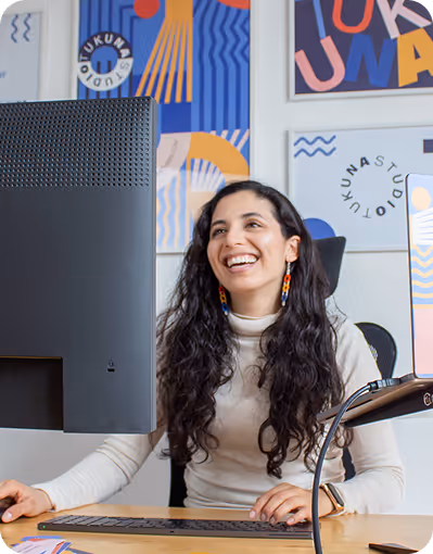 Smiling woman with long curly hair sitting at a desk working on a computer in a colorful art-filled office.