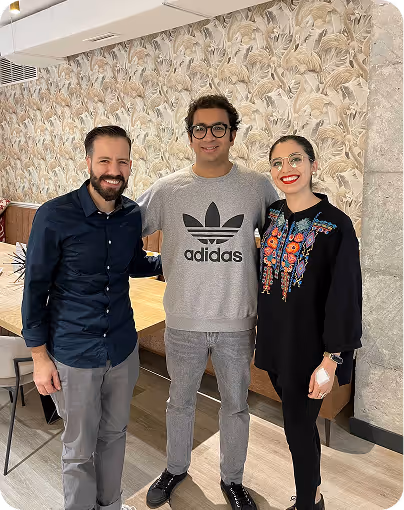 Three people standing indoors smiling, one wearing an Adidas sweatshirt and glasses.