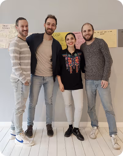 Four casually dressed people standing closely indoors, smiling in front of a wall with colorful sticky notes.