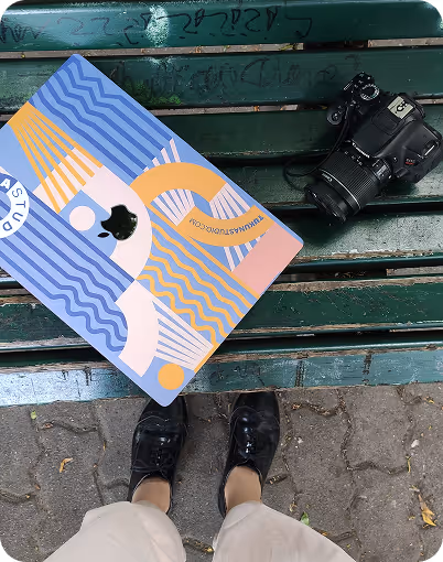 Colorful laptop with abstract design, DSLR camera, and a person’s feet on green wooden bench outdoors.