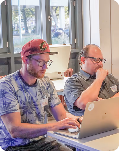 Two men wearing glasses sitting at a table working on laptops in a well-lit room.
