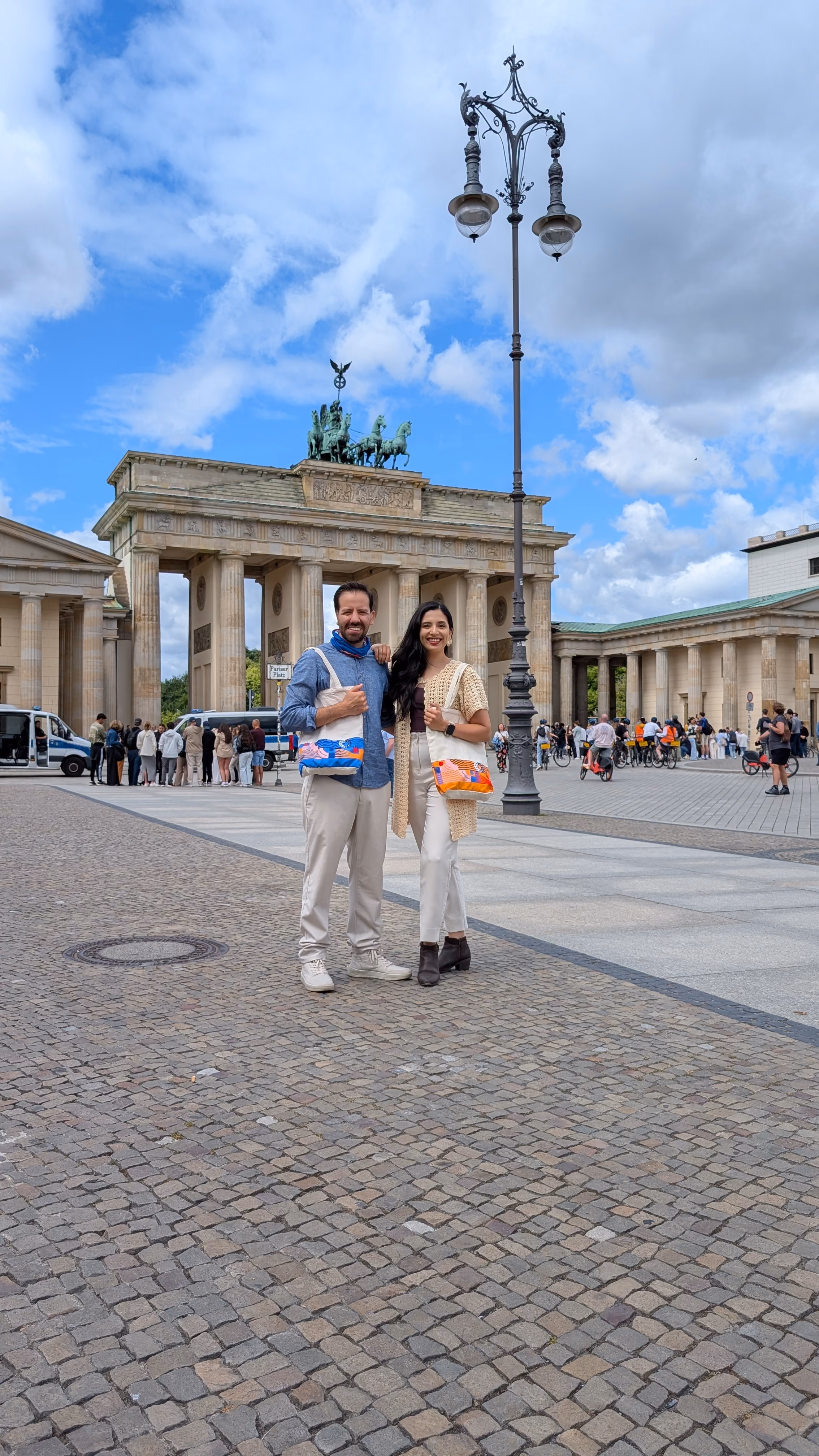 Smiling man and woman holding colorful paper shapes in front of the Brandenburg Gate in Berlin.
