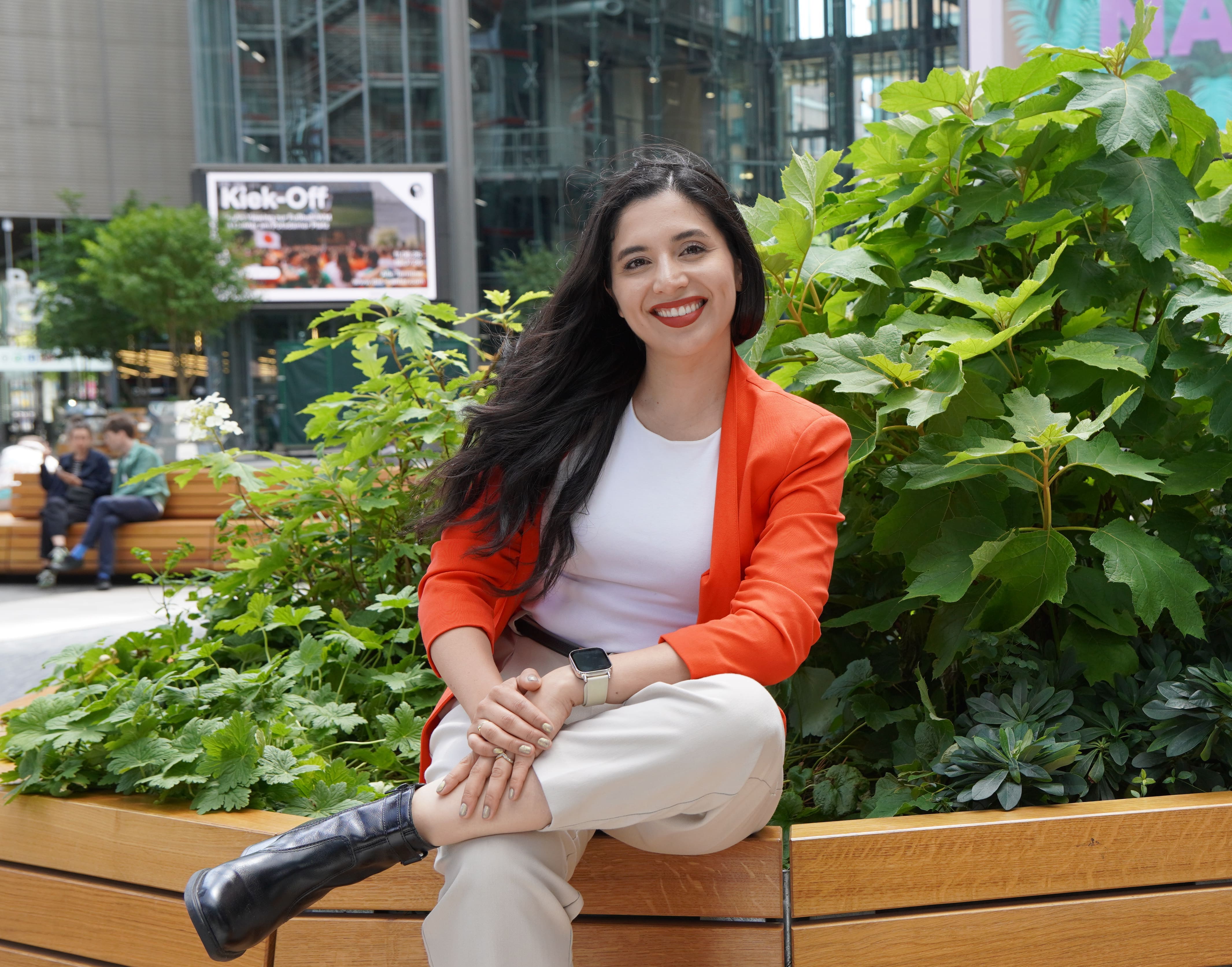 Smiling woman with dark hair and red lipstick standing in front of colorful abstract wall art and green plants.