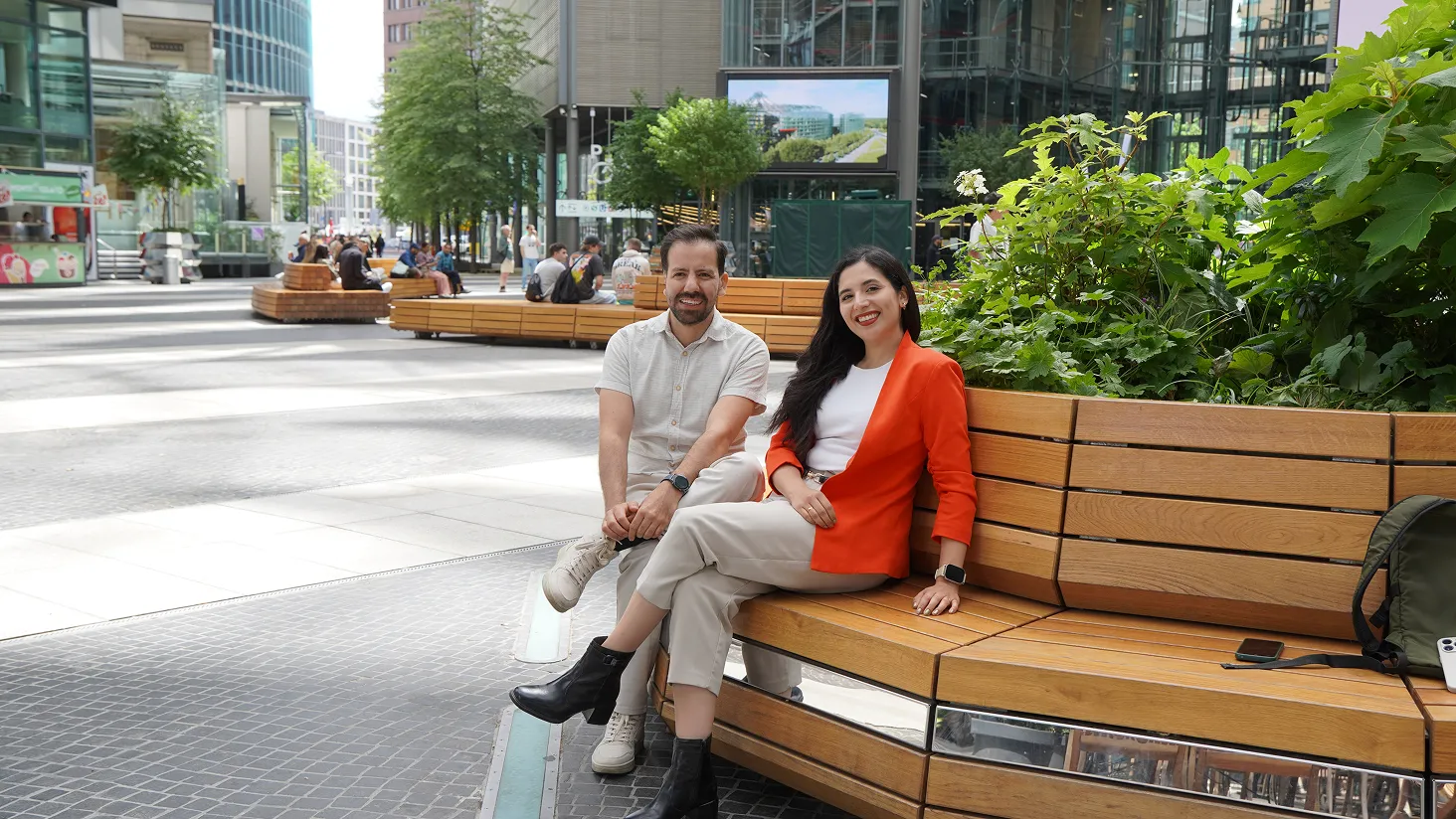 Man and woman sitting and smiling on a wooden bench surrounded by plants in an urban plaza.