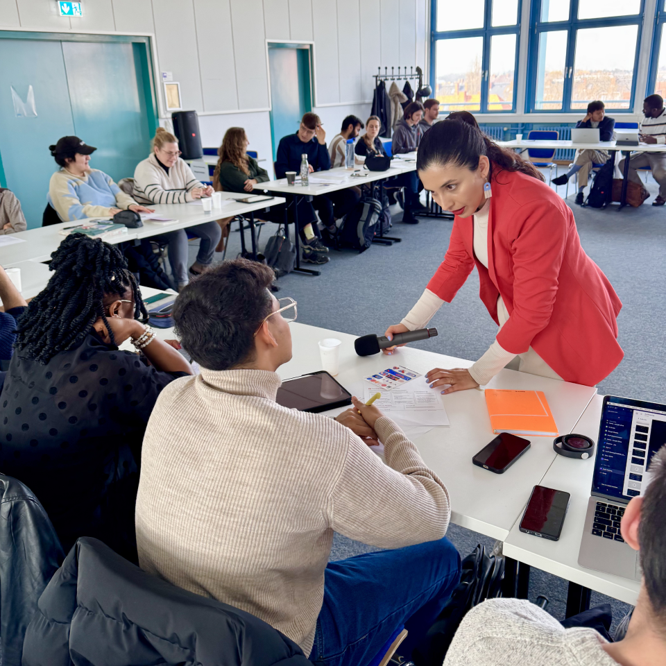 Woman in red blazer holding microphone to man speaking during a group discussion in a classroom.
