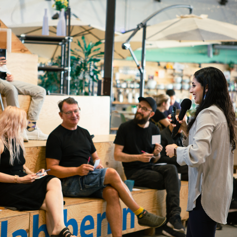 Woman speaking with microphone to seated audience in casual indoor event space with plants and umbrellas.