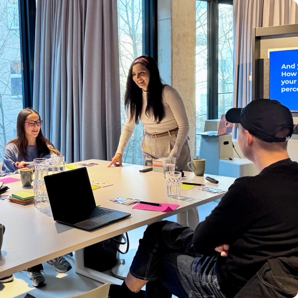 Three people engaged in a bright meeting room with laptops, notes, and a screen in the background.