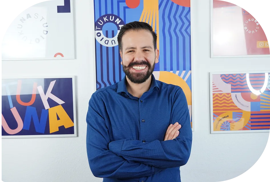 Man with a beard and mustache wearing a blue shirt, smiling in front of colorful framed artwork on a white wall.