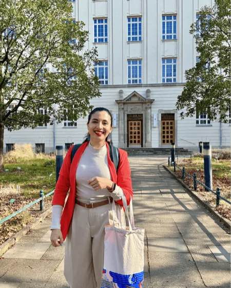 Woman in red blazer and beige pants smiling, standing on a path in front of a large white building with windows.