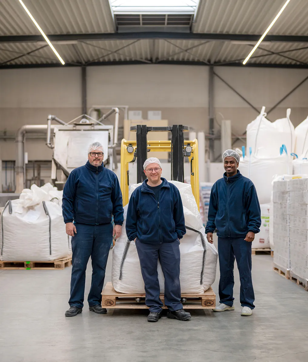 Drie werknemers van Caribbean Gold in blauwe overalls en haarnetjes staan in een magazijn naast een pallet met grote witte zakken en een gele heftruck.