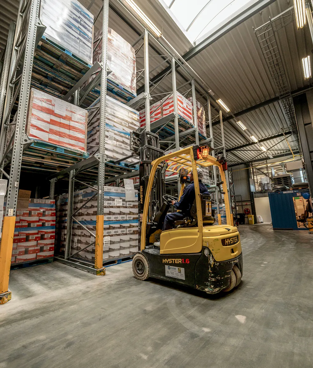 Magazijn van Caribbean Gold met hoge stellingen gevuld met dozen, waar een heftruckchauffeur met een gele Hyster-heftruck pallets verplaatst.