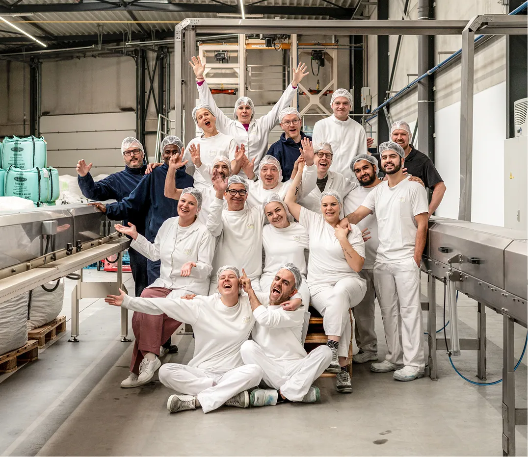 Het team van Caribbean Gold in witte en blauwe bedrijfskleding en harenbescherming die vrolijk in een productieomgeving poseert.