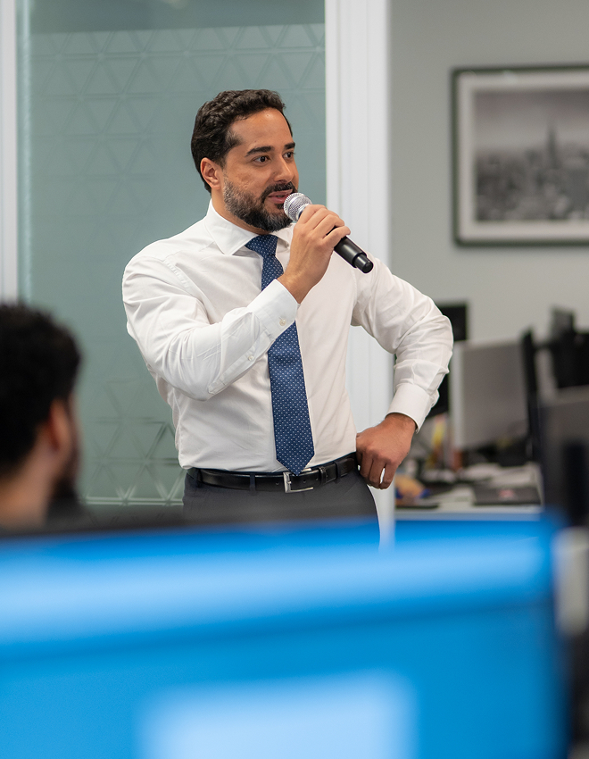 Homem vestido com camisa branca e gravata azul falando ao microfone em um escritório moderno.