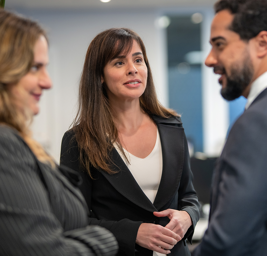 Três profissionais em ambiente de escritório, uma mulher ao centro falando com outra mulher e um homem em traje formal.