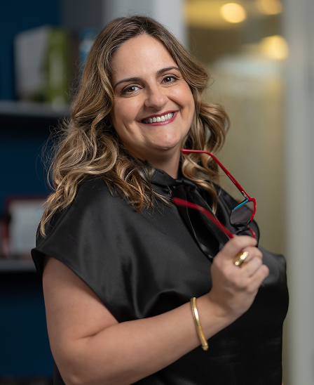 Mulher sorridente de cabelo ondulado segurando óculos de armação vermelha, vestindo blusa preta, em ambiente interno desfocado.