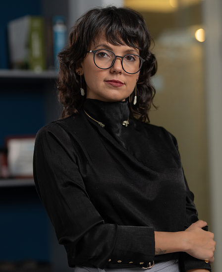 Mulher jovem com cabelo cacheado curto e óculos redondos, vestindo blusa preta e brincos longos, olhando para a câmera com expressão confiante.