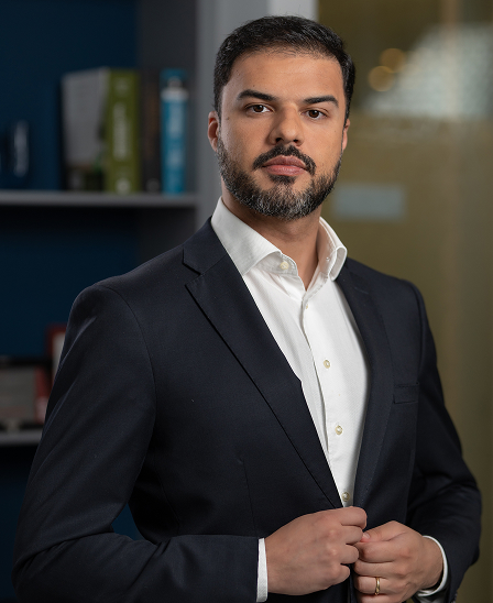 Homem de barba e cabelo curto vestindo terno preto e camisa branca, ajustando o paletó em um ambiente interno com estante ao fundo.