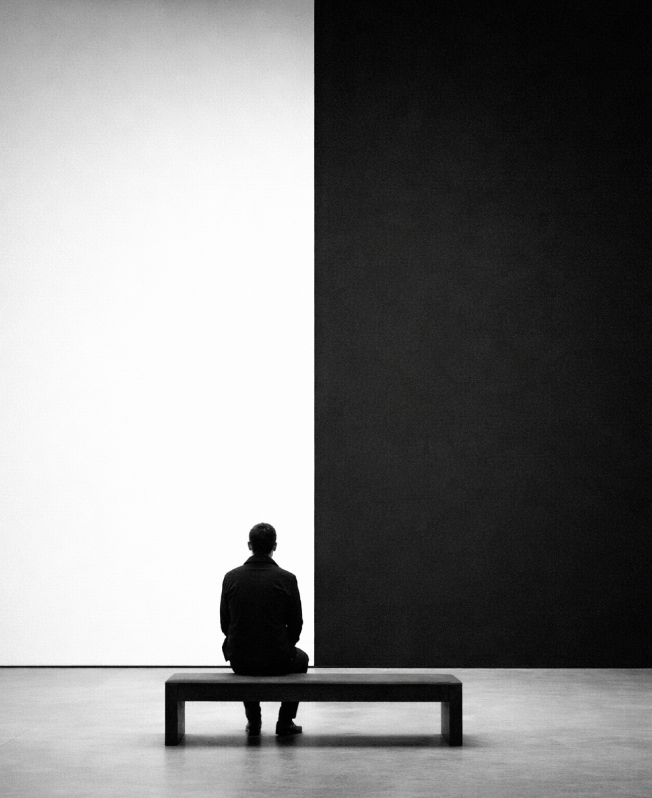 Pessoa sentada de costas em um banco diante de uma parede dividida em preto e branco.