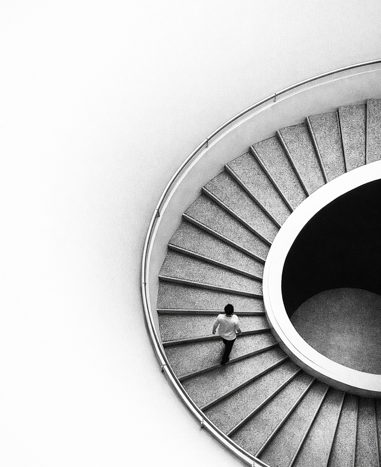 Pessoa descendo uma escada espiral cinza vista de cima em uma imagem em preto e branco.