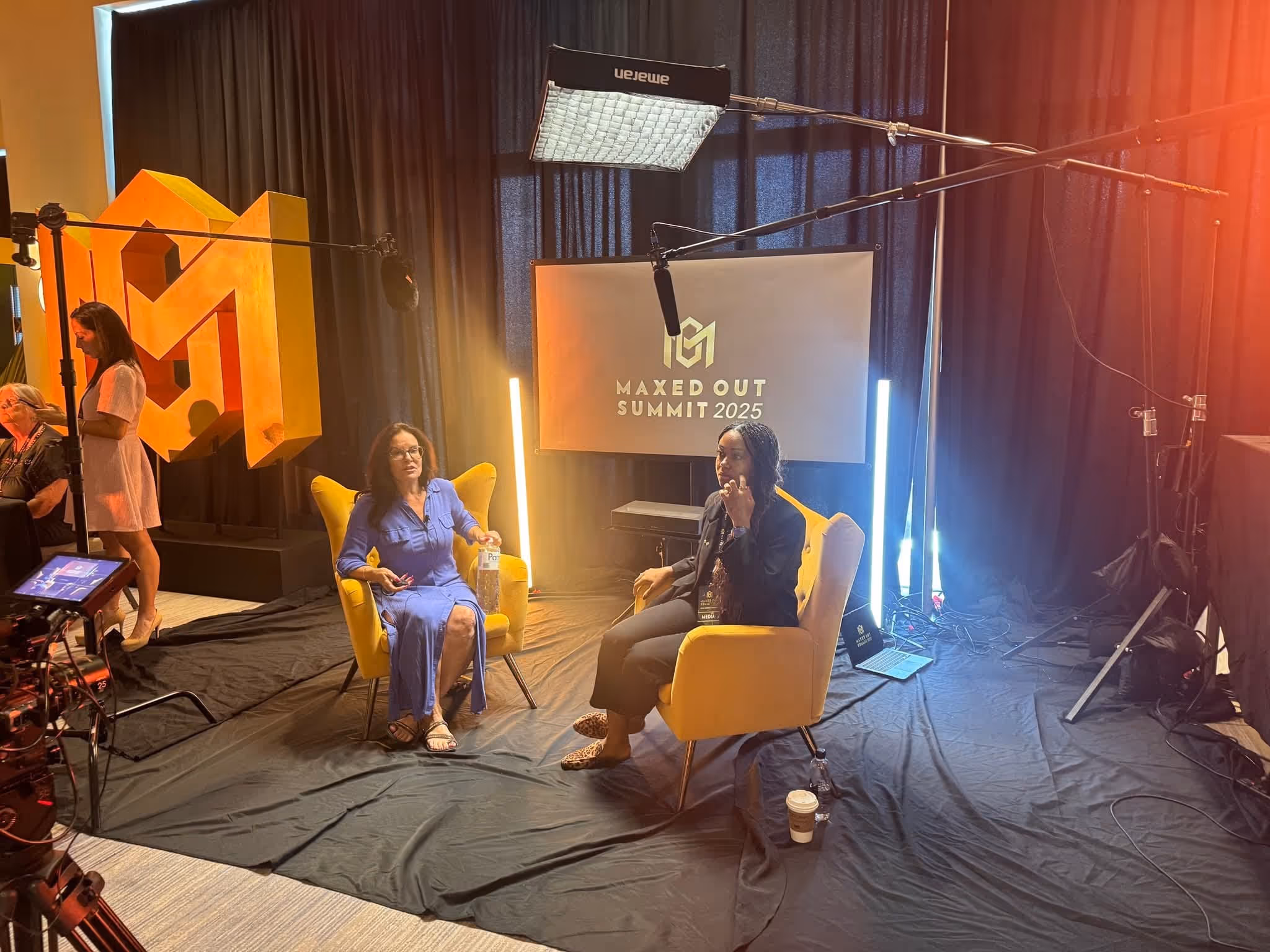 Two women seated in yellow armchairs during an interview setup with lighting, microphones, and a 'Maxed Out Summit 2025' sign in the background.