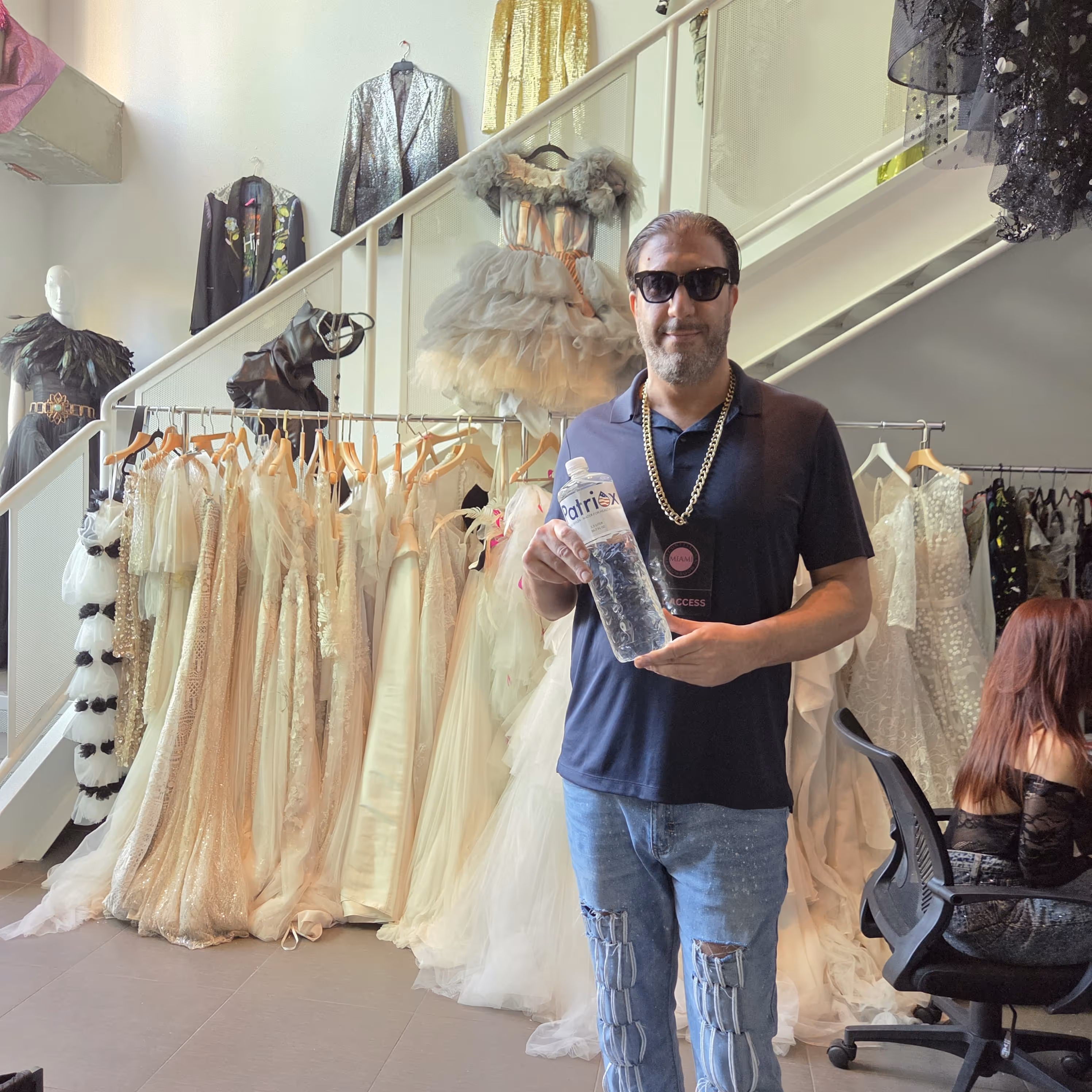 Man wearing sunglasses and a gold chain holding a large water bottle in front of a display of elegant white dresses and gowns in a fashion studio.