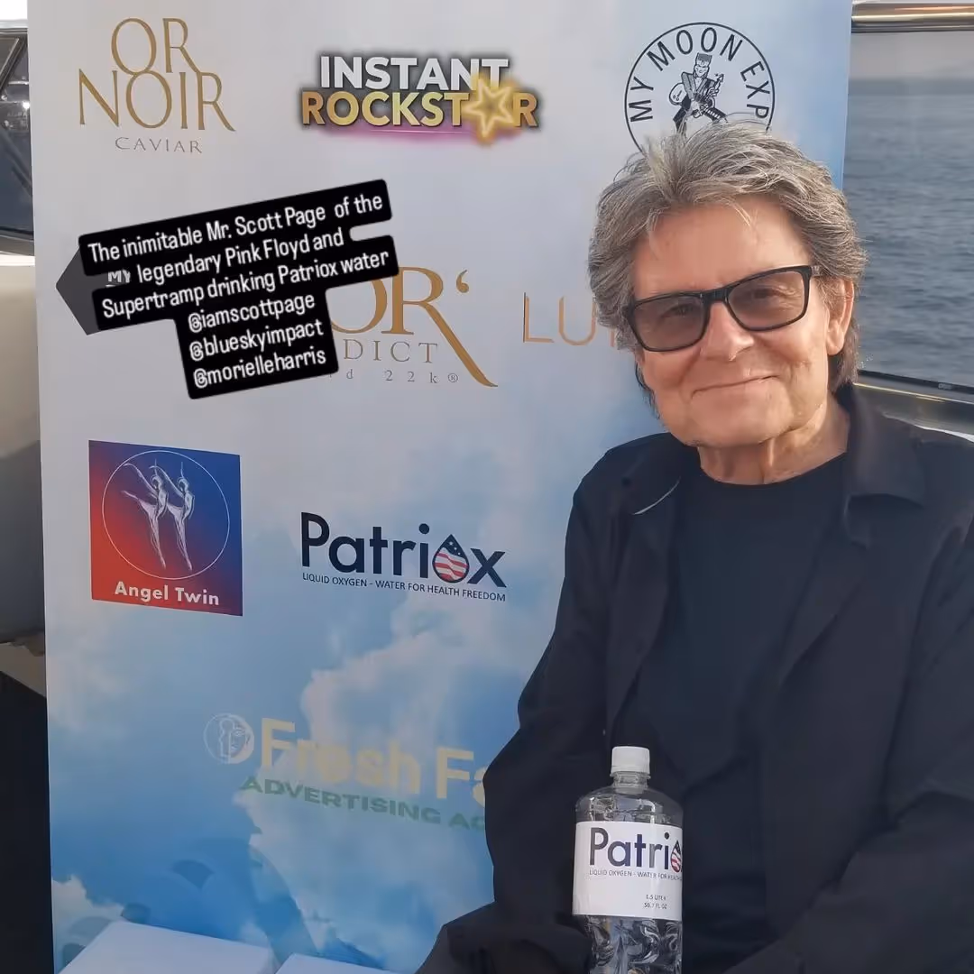 Man with gray hair and glasses holding a bottle of Patriox water, sitting in front of a promotional backdrop with various logos.