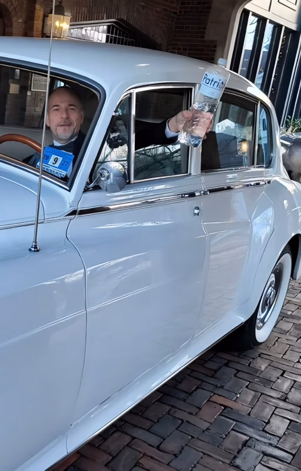 Man in a white vintage car holding a water bottle out of the passenger window on a brick driveway.
