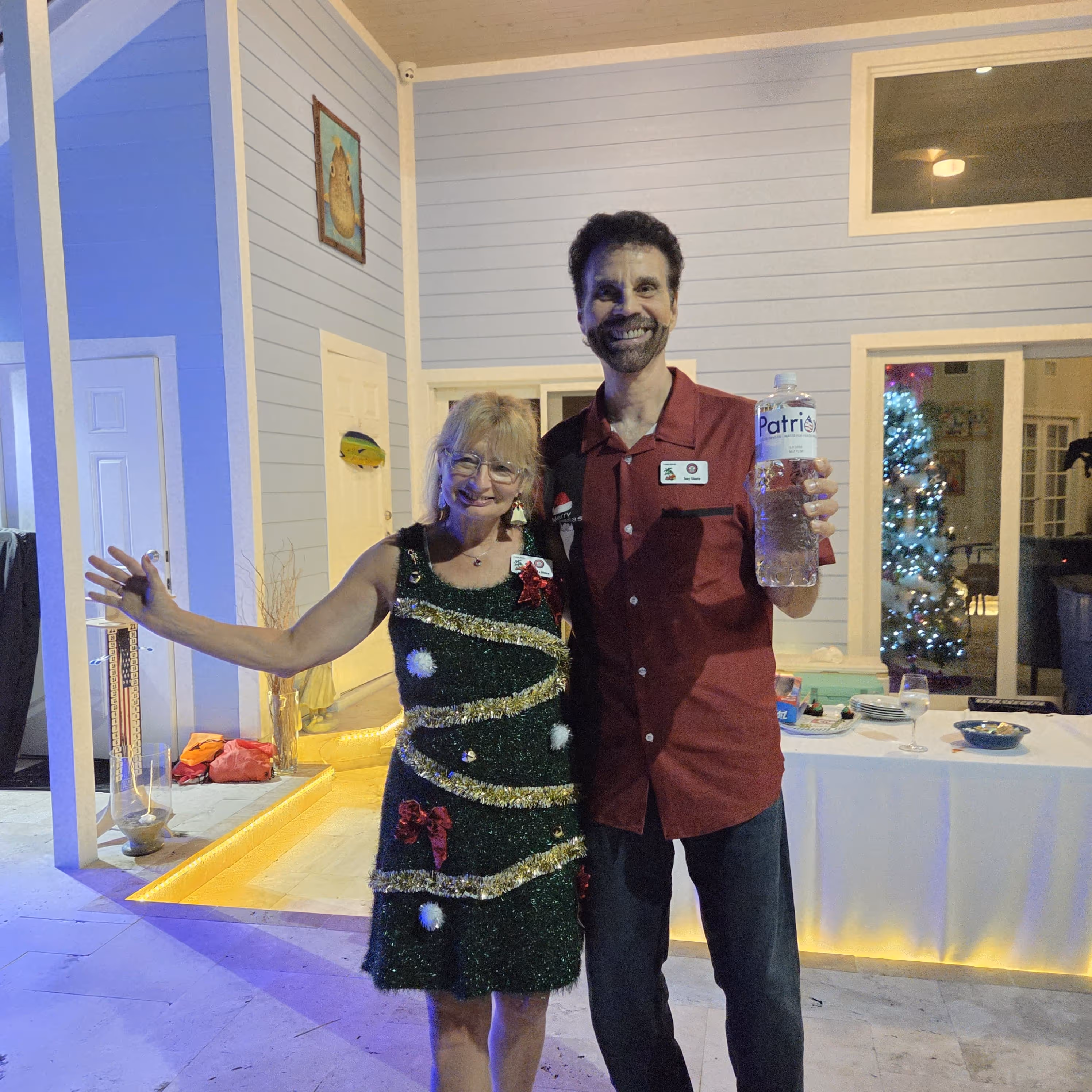 Smiling man and woman standing indoors near decorated Christmas tree, woman wearing a festive dress and man holding a water bottle.