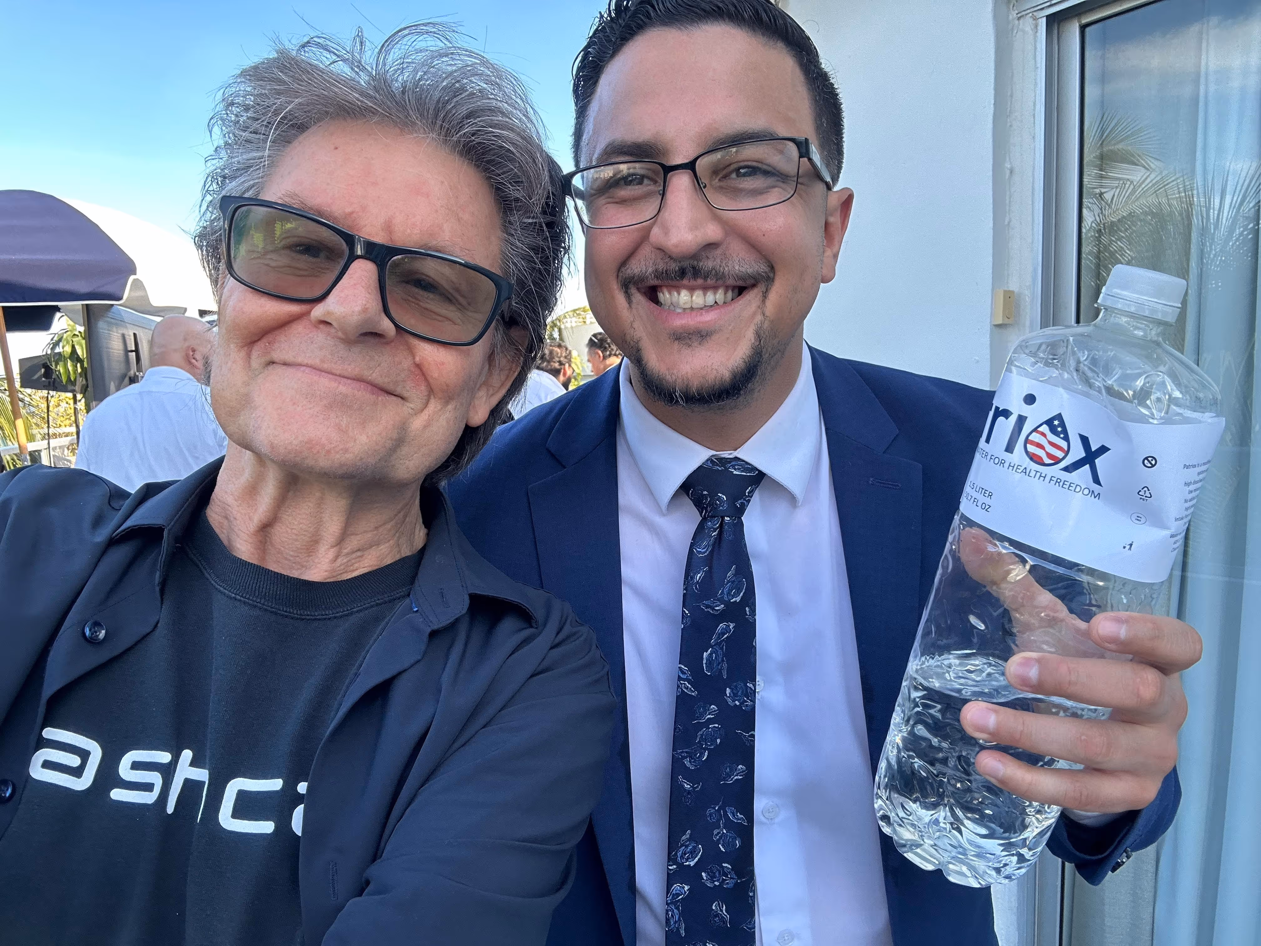 Two men smiling for a selfie, one wearing glasses and a black shirt and jacket, the other in a blue suit and tie holding a large water bottle labeled 'riox'.