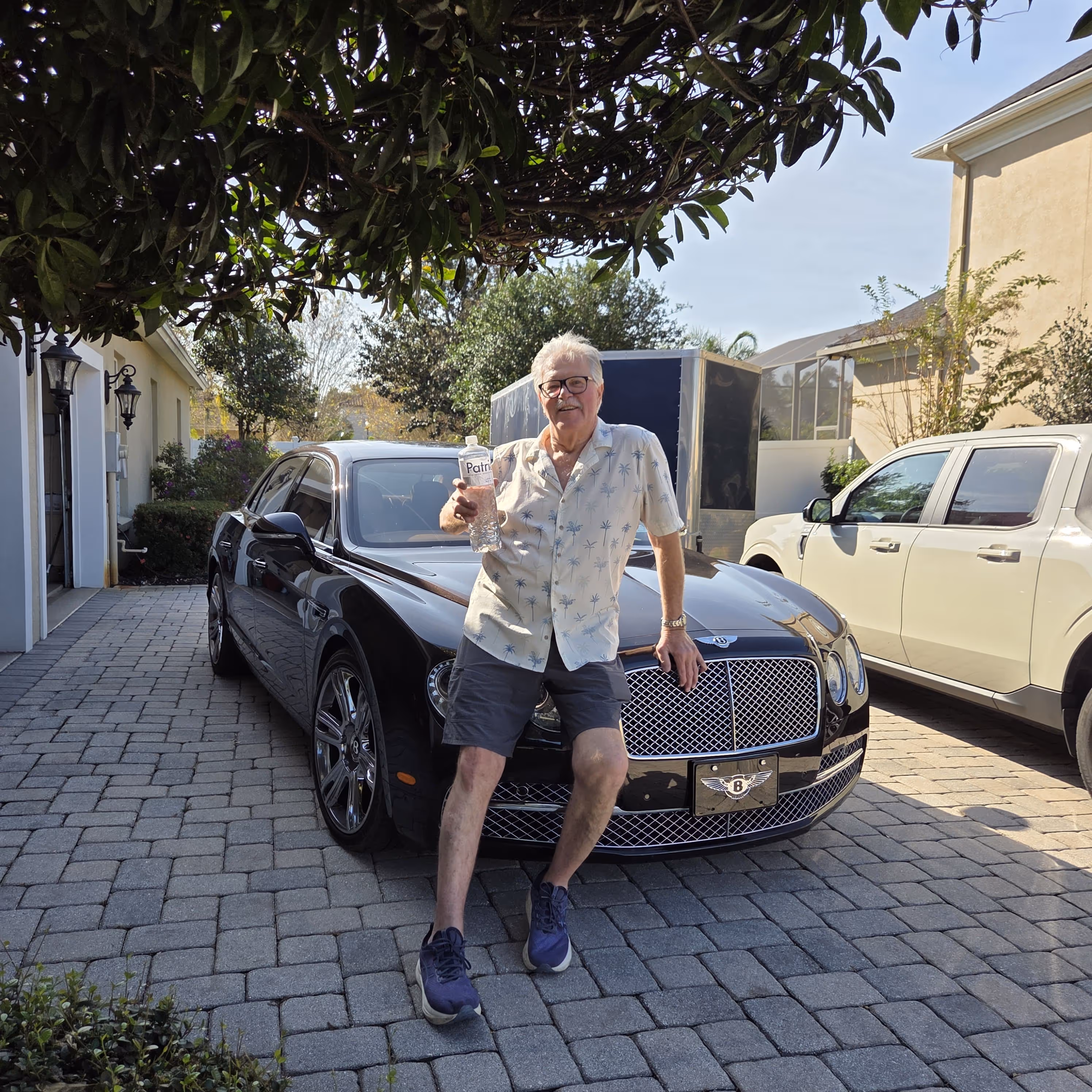 Older man in a palm tree print shirt and shorts sitting on the front of a black Bentley parked on a driveway, holding a bottle of water.