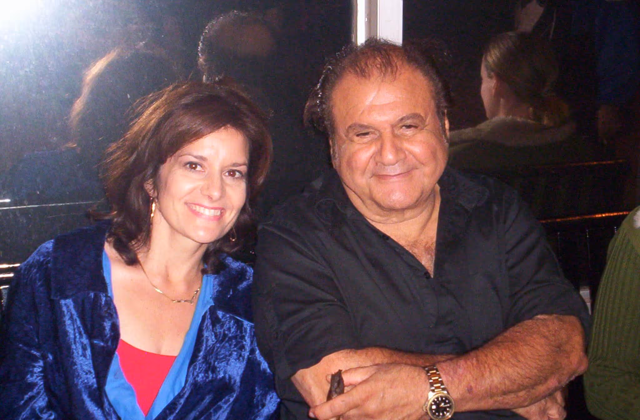 Smiling woman in blue velvet jacket sitting beside a man in a black shirt with crossed arms.