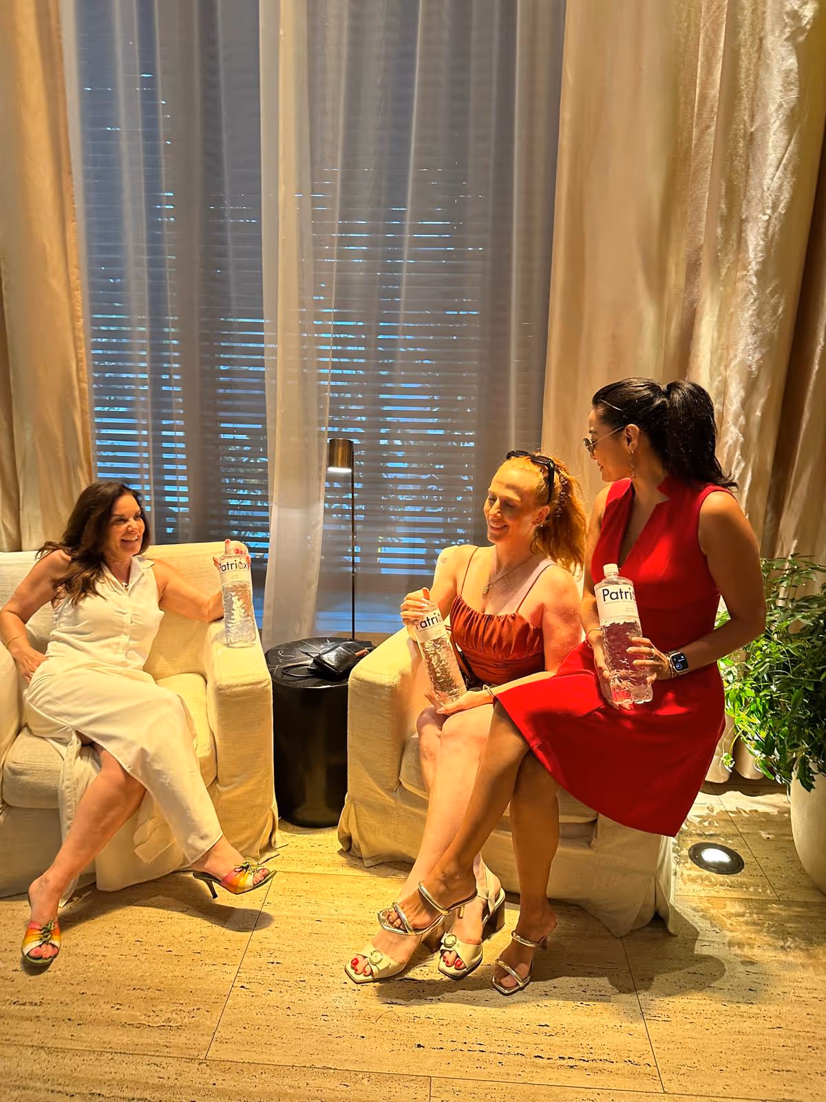 Three women sitting on beige armchairs indoors, smiling and holding bottles of water labeled 'Patria'.