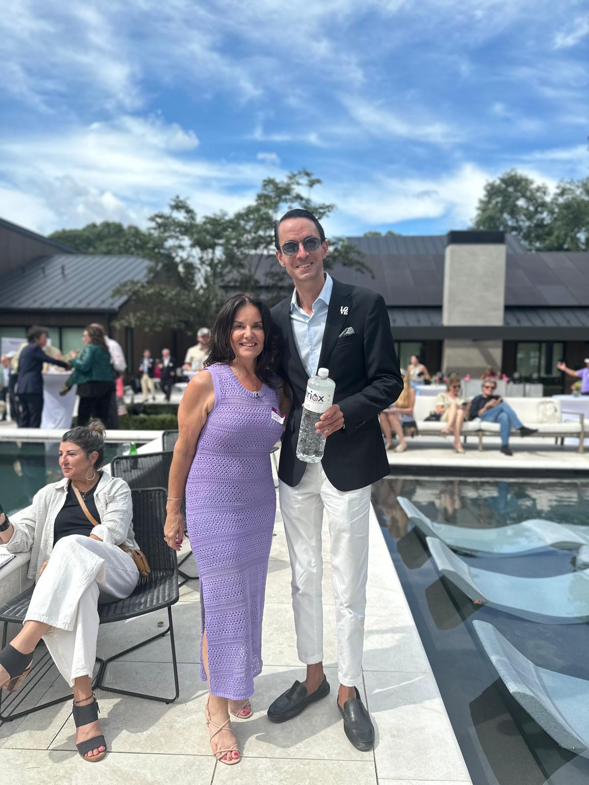 A smiling man in a black blazer and white pants holding a water bottle stands next to a woman in a lavender sleeveless dress beside an outdoor pool with lounge chairs.