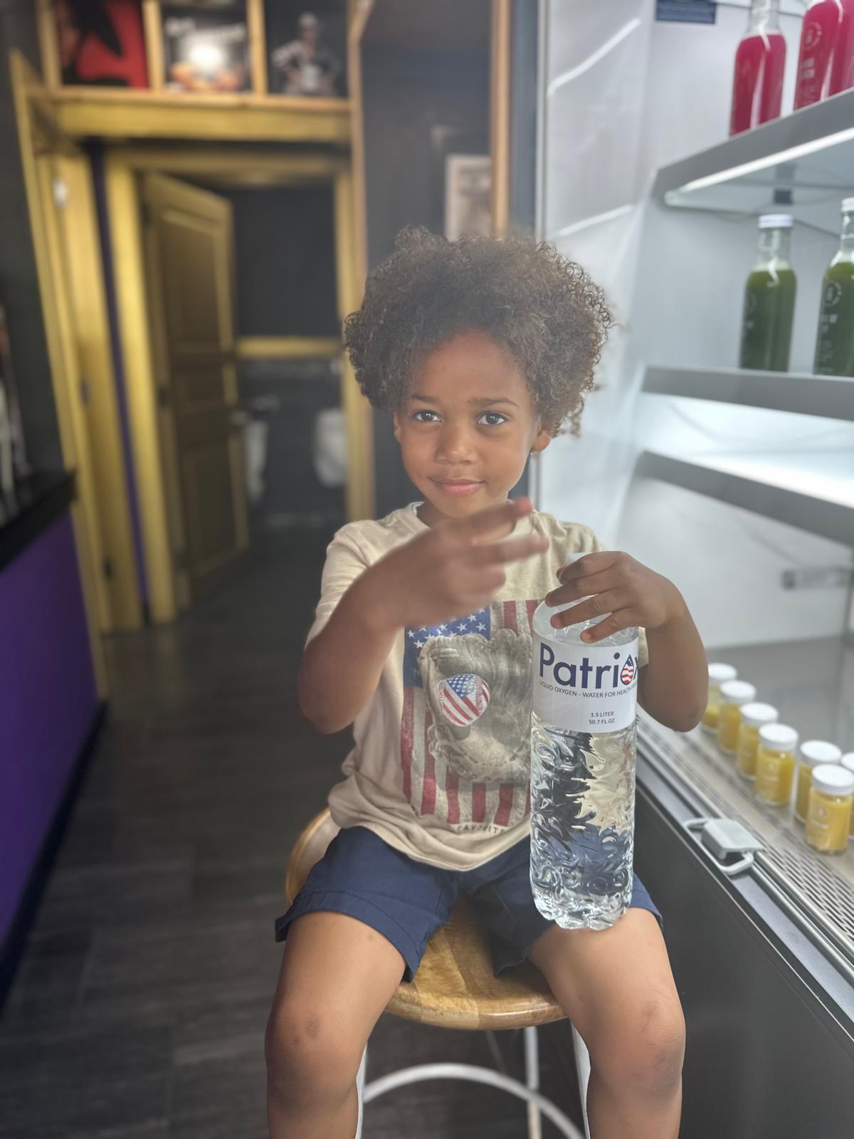 Young child with curly hair sitting on a wooden stool, holding a large bottle of Patri water.