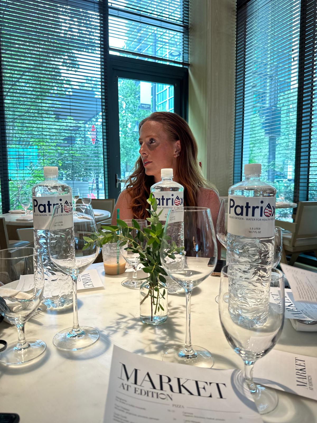 Woman with long hair sitting at a restaurant table with three bottles of Patriox water, wine glasses, a small vase with green leaves, and a Market at Edition menu.