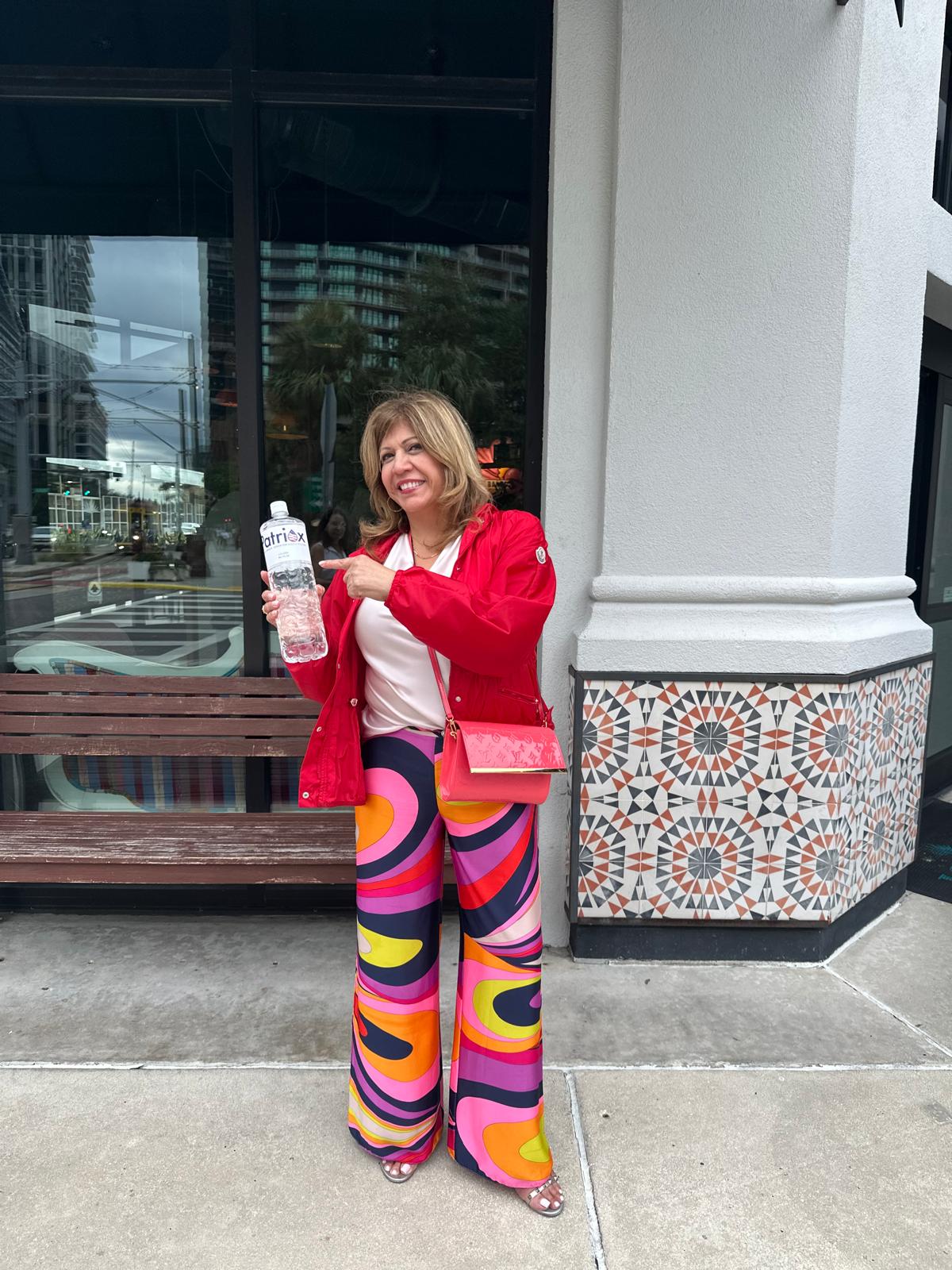 Woman in a red jacket and colorful patterned pants pointing to a bottle of water while standing on a city sidewalk.