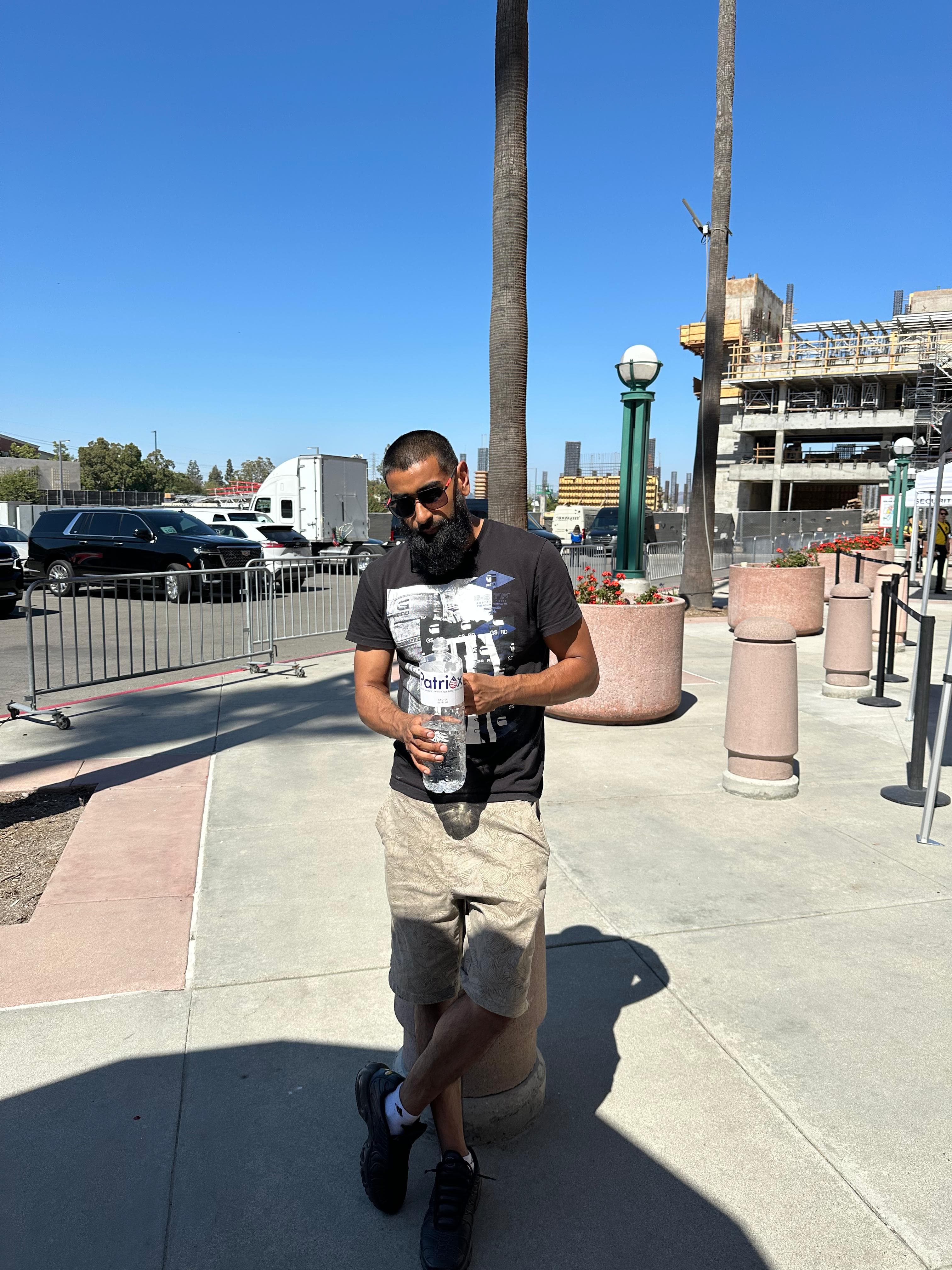 Bearded man wearing sunglasses, black graphic t-shirt, and beige shorts holding a water bottle and leaning against a concrete post outdoors near parking and construction site.