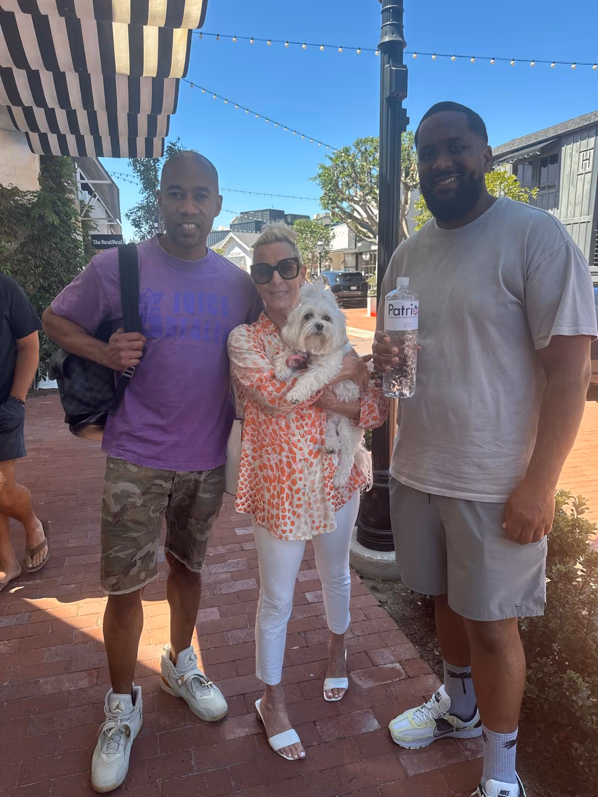 Three people standing outdoors on a brick sidewalk under string lights, one woman holding a small white dog, one man wearing a purple shirt and camo shorts, and another man holding a bottle of water.
