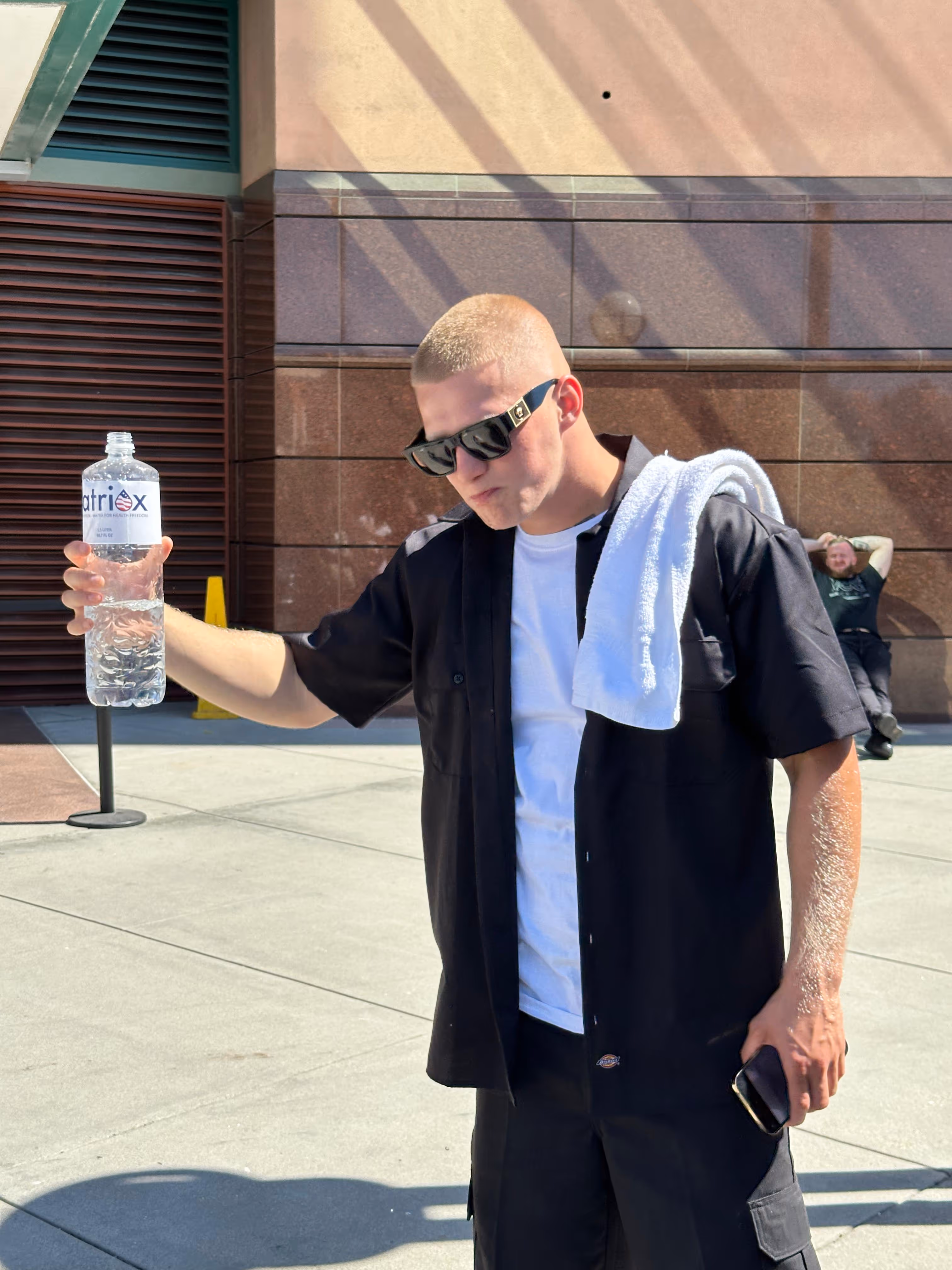 Man wearing sunglasses, a black shirt over a white t-shirt, with a white towel around his neck, holding a large water bottle outdoors on a sunny day.